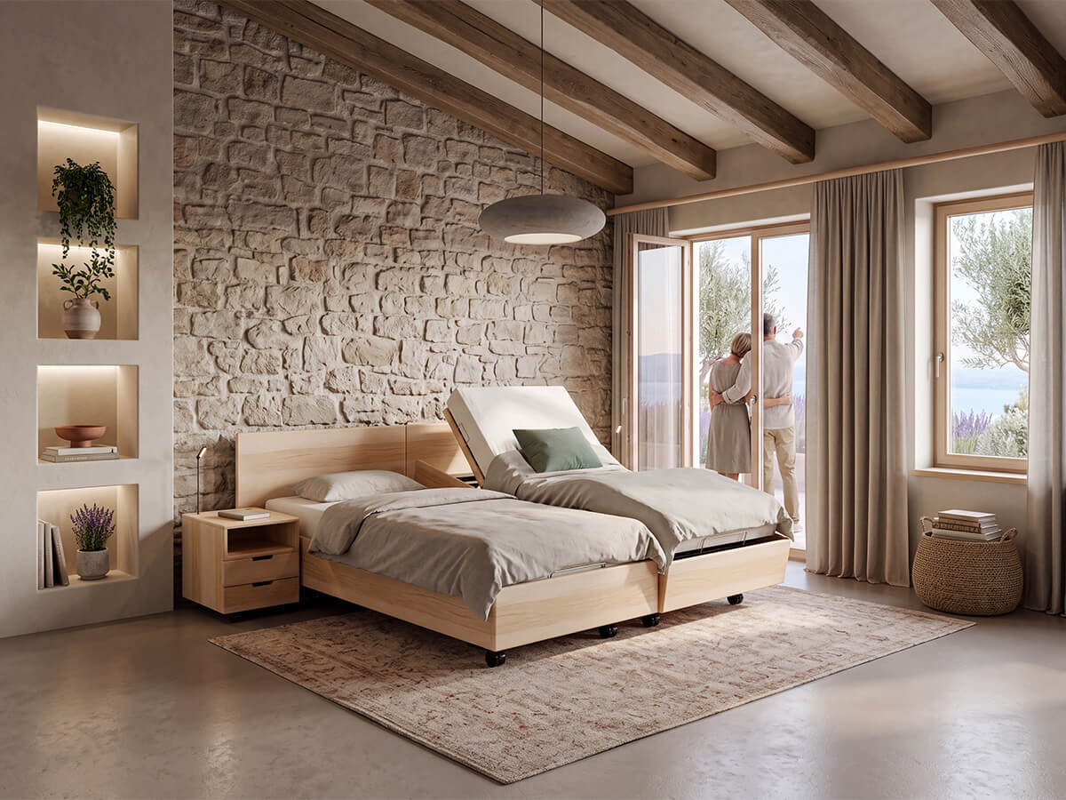 Massivholzbett Bergen Classic von KIRCHNER in Eiche bianco mit hochgestelltem Kopfteil auf einer Seite in einem Schlafzimmer mit Holzbalkendecke und Natursteinwand, ein Paar steht umarmt am Fenster