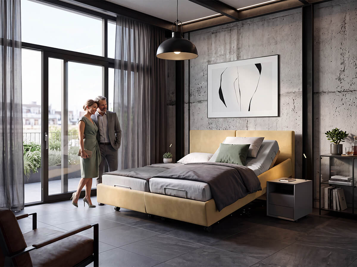 Polsterbett Nevara Classic von KIRCHNER mit senfgelbem Polster, hochgestelltem Kopfteil und geneigtem Bettrahmen auf einer Seite in einem modernen Schlafzimmer mit Betonwänden bei Tag, ein Paar steht am Fenster