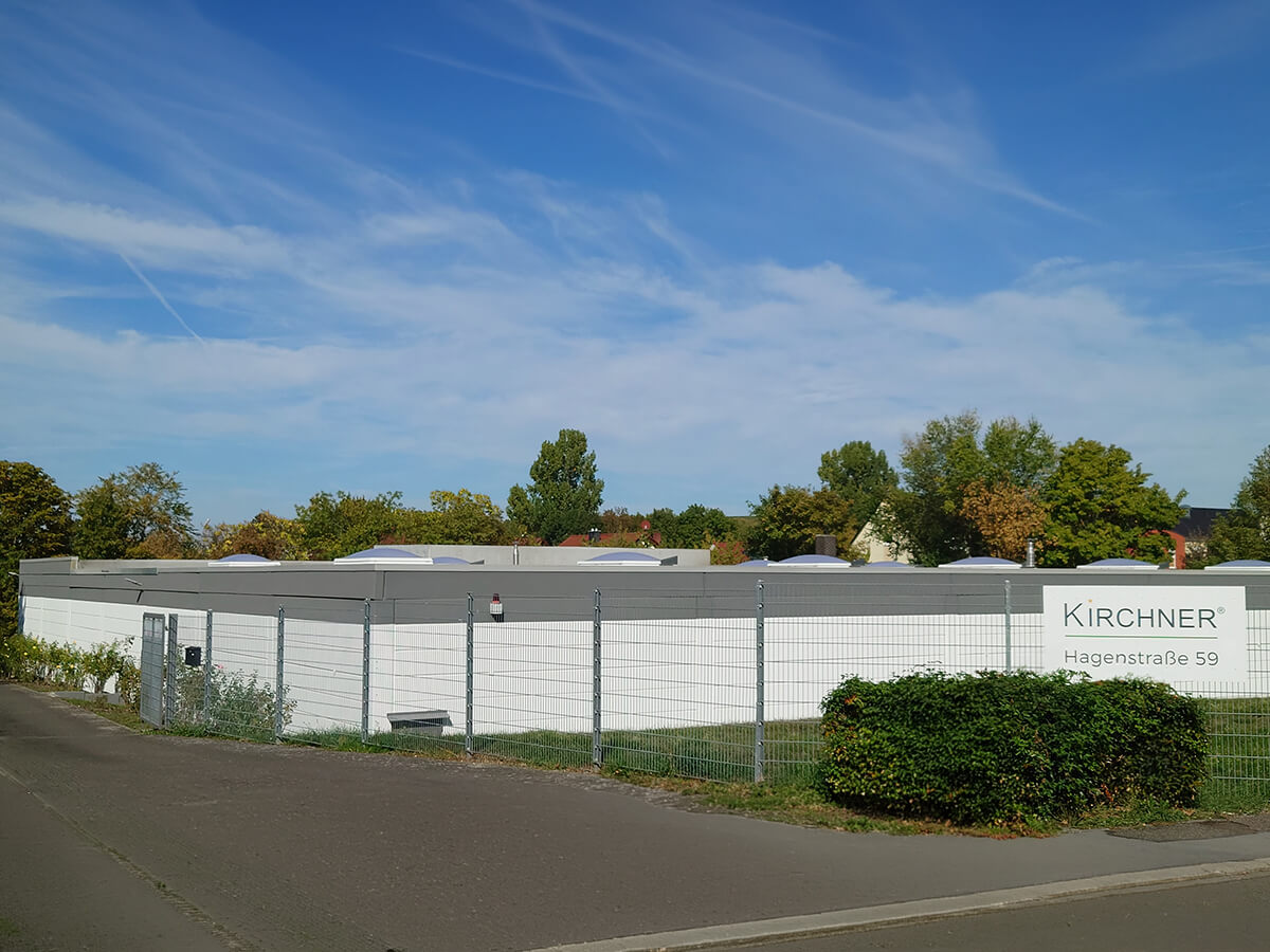 Außenansicht des KIRCHNER®-Unternehmensstandorts in Alzey mit Firmengebäude, Einfahrt, Zaun und Firmenschild.