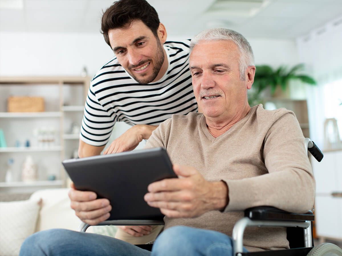 Älterer Mann im Rollstuhl checkt zusammen mit seinem Sohn am Tablet den Bettenkonfigurator. 