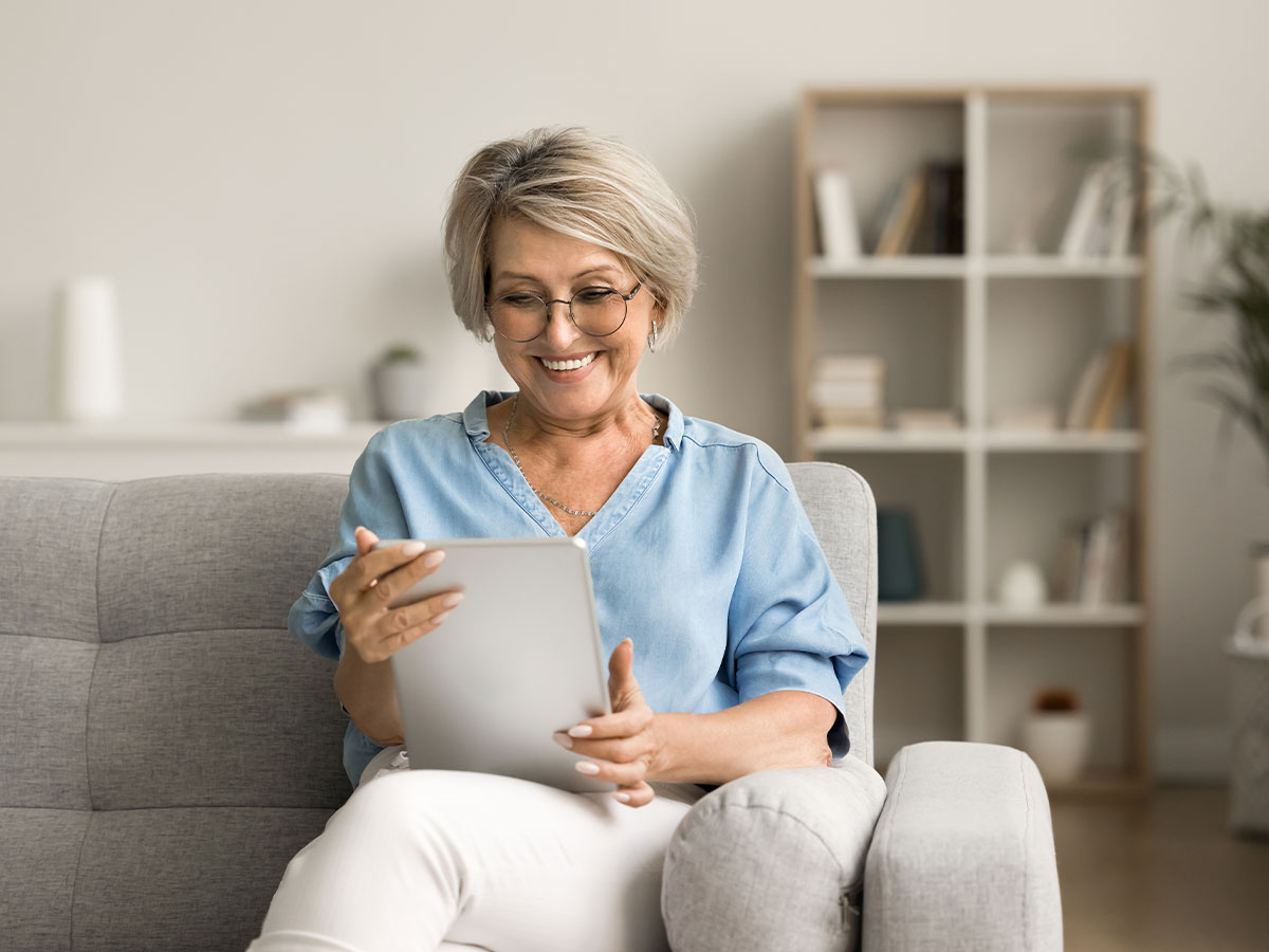 Eine ältere Frau mit Brille sitzt zu Hause auf dem Sofa und guckt in ihr Tablet