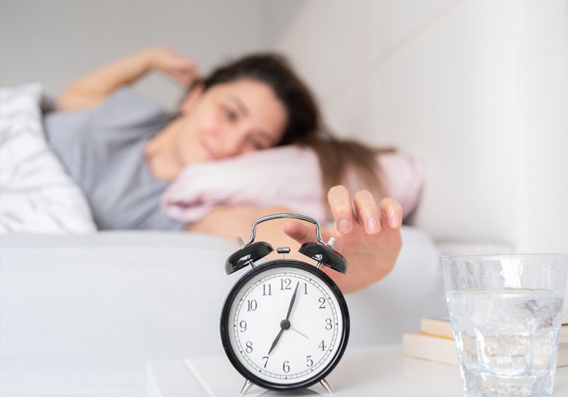 Eine junge Frau wacht lächelnd in ihrem Bett auf und greift nach ihrem Wecker