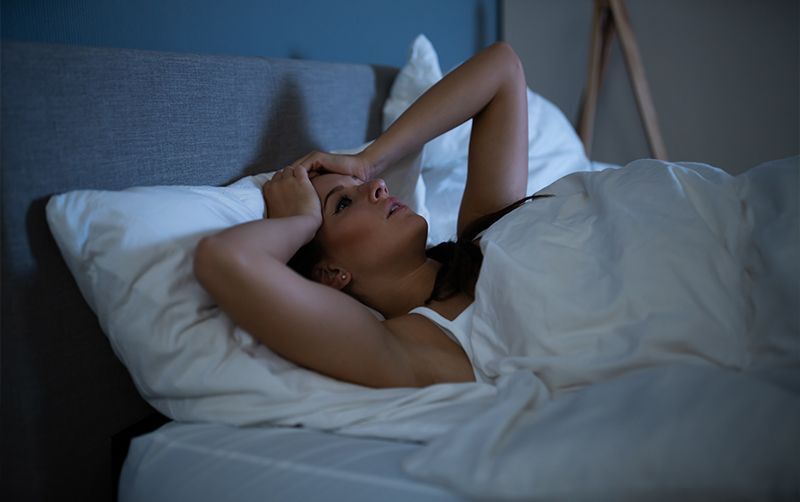 Frau liegt auf dem Bett und hält sich die Hände an den Kopf, weil sie nicht einschlafen kann.