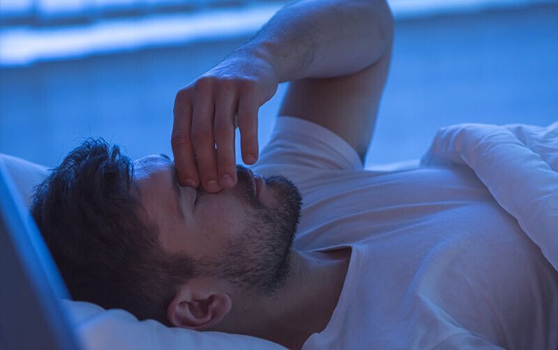 Mann, der in den Morgenstunden wach im Bett liegt und sich die linke Hand auf den Nasenrücken drückt