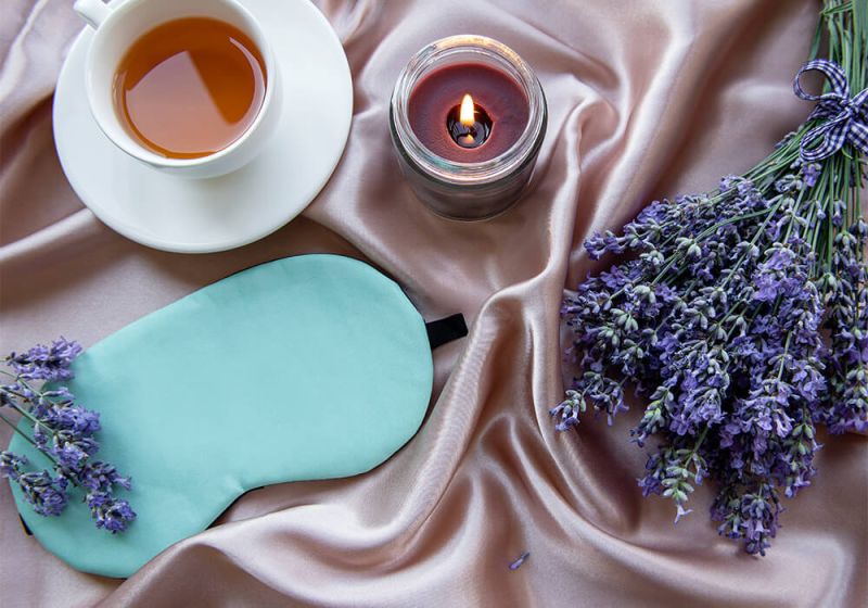 Getrockneter Lavendel, eine Tasse Tee, eine Schlafmaske sowie eine brennende Kerze auf einer rosafarbenen Bettdecke.