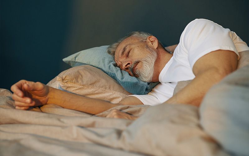 Ein Mann mit grauen Haaren und Bart liegt bei optimaler Schlafhygiene entspannt im Bett und schläft.