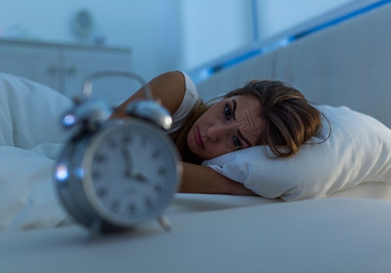 Frau mit Schlafproblemen liegt wach in ihrem Bett und starrt auf einen Wecker